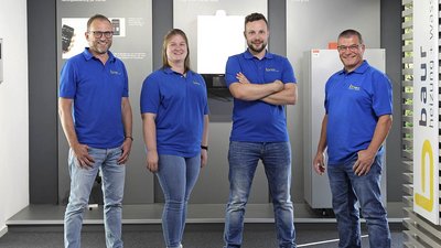 Vier Personen in blauen Poloshirts stehen lächelnd in einem modernen Ausstellungsraum mit Heizungsgeräten im Hintergrund.