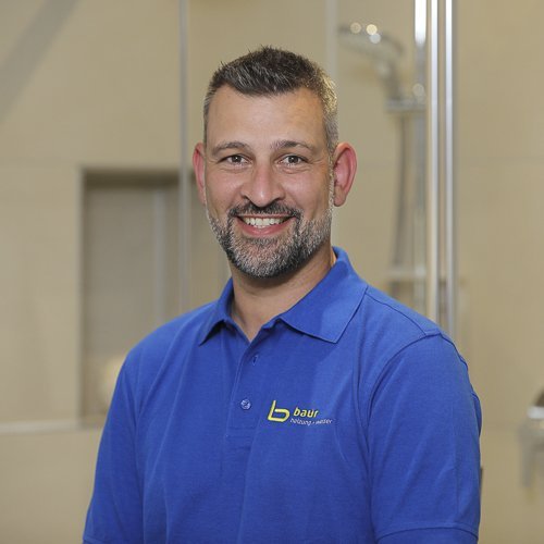 Ein lächelnder Mann mit kurzem Bart trägt ein blaues Poloshirt und steht vor einer hellen, modernen Umgebung.