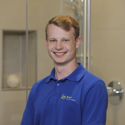 Ein junger Mann mit blonden Haaren lächelt freundlich in einem modernen Badezimmer mit Glasduschkabine im Hintergrund.