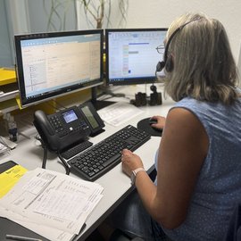Eine Frau mit grauen Haaren sitzt an einem Schreibtisch und arbeitet konzentriert an zwei Monitoren.