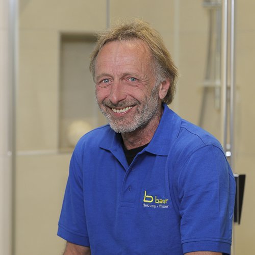 Ein lächelnder Mann mit grauen Haaren trägt ein blaues Polo-Shirt und sitzt in einem hellen, modernen Badezimmer.