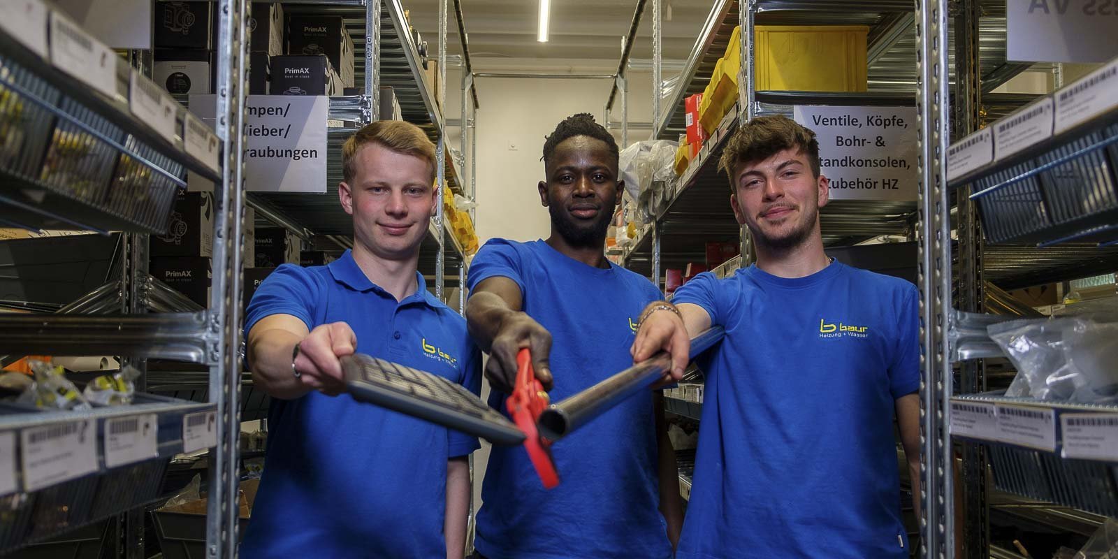 Drei Männer in blauen T-Shirts stehen in einem Lager und halten Werkzeuge in der Hand, lächelnd und bereit zur Arbeit.