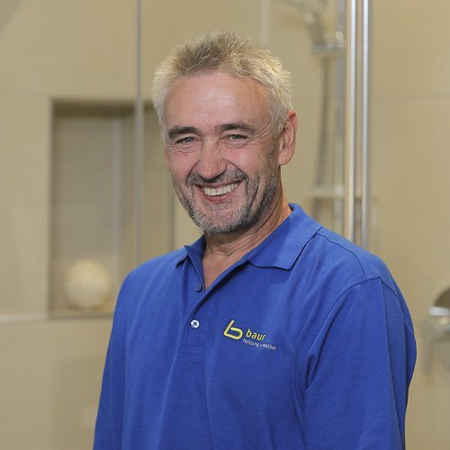 Ein lächelnder Mann mit grauen Haaren trägt ein blaues Polohemd und steht in einem modernen Badezimmer.