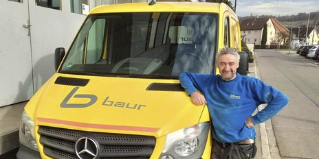 Ein lächelnder Mann in blauer Kleidung steht entspannt neben einem gelben Lieferwagen mit dem Logo 'baur'.