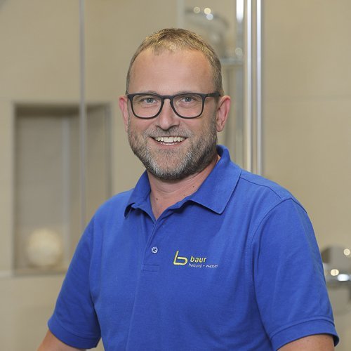 Ein lächelnder Mann mit Brille trägt ein blaues Polohemd und steht in einem modernen Badezimmer mit hellen Fliesen.