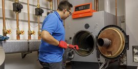 Ein Techniker in blauer Uniform arbeitet an einer großen Heizungsanlage und benutzt ein Werkzeug zur Wartung.