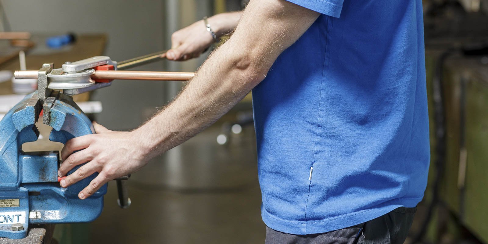 Ein Handwerker in einem blauen T-Shirt arbeitet mit einer Rohrzange an einem Kupferrohr in einer Werkstatt.