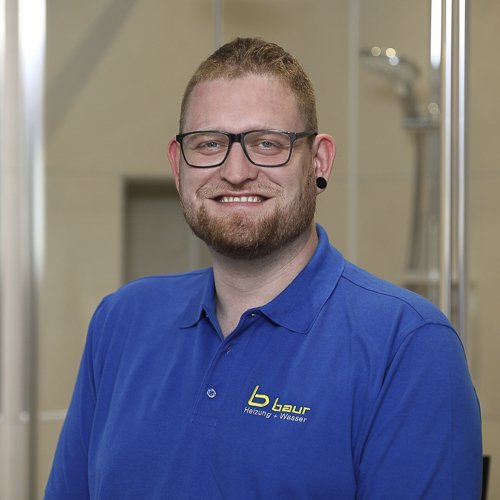 Ein lächelnder Mann mit Brille trägt ein blaues Polohemd und steht vor einer modernen, hellen Umgebung.