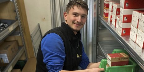 Ein junger Mann lächelt, während er eine grüne Kiste mit Werkzeugen in einem Lagerregal sortiert.