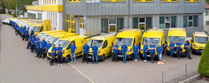 Eine Gruppe von Mitarbeitern in blauen Shirts steht lächelnd vor einer Reihe gelber Lieferwagen in einem Firmenhof.