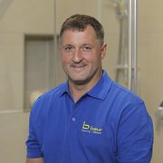 Professioneller Handwerker in blauer Arbeitskleidung lächelt freundlich Ein lächelnder Mann in einem blauen Poloshirt steht vor einer modernen Badezimmerumgebung mit Glasduschkabine.