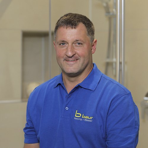 Ein lächelnder Mann in einem blauen Poloshirt steht vor einer modernen Badezimmerumgebung mit Glasduschkabine.
