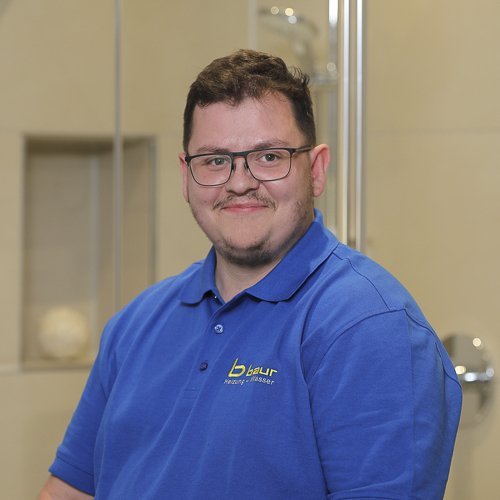 Ein lächelnder Mann mit Brille trägt ein blaues Polohemd und sitzt in einem modernen Badezimmer mit hellen Fliesen.