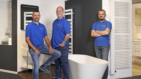 Drei Männer in blauen T-Shirts stehen in einem modernen Badezimmerausstellungsraum, umgeben von Sanitäranlagen.