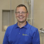 Freundlicher Mitarbeiter präsentiert Badezimmerlösungen mit Begeisterung Der Mann mit Brille und einem breiten Lächeln trägt ein Poloshirt mit dem Logo von Baur, umgeben von hellen Fliesen.