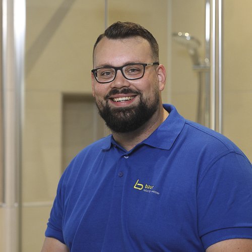 Ein lächelnder Mann mit Brille trägt ein blaues Poloshirt und steht in einem modernen Badezimmer mit Glasduschkabine.