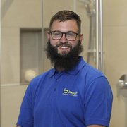 Professioneller Handwerker in blauer Arbeitskleidung lächelt freundlich Ein lächelnder Mann mit Bart trägt ein blaues Poloshirt und steht in einem modernen Badezimmer mit Glasduschkabine.