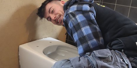 Ein junger Mann mit einem Lächeln beugt sich über eine Toilette in einem Raum mit beige Wänden und dunklen Fliesen.