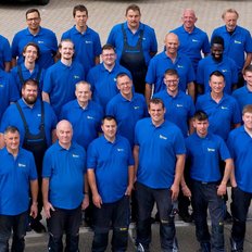 Männer in blauen Poloshirts posieren gemeinsam auf einem Parkplatz, umgeben von Autos und einem gelben Lieferfahrzeug.