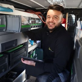 In einem Lieferwagen sortiert ein Handwerker seine Werkzeuge und schaut freundlich in die Kamera.