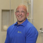 Lächelnder Mann in blauer Arbeitskleidung im Badezimmer Ein lächelnder Mann in einem blauen Poloshirt steht in einem modernen Badezimmer mit hellen Fliesen und einer Dusche im Hintergrund.