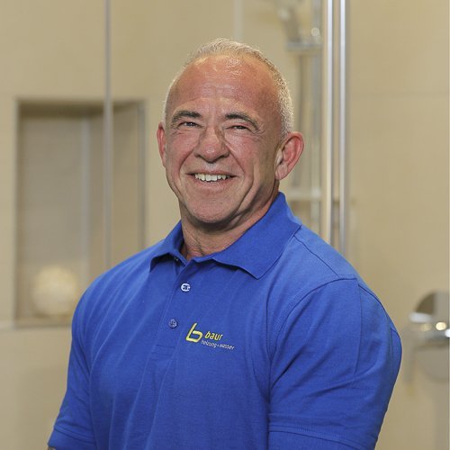 Ein lächelnder Mann in einem blauen Poloshirt steht in einem modernen Badezimmer mit hellen Fliesen und einer Dusche im Hintergrund.