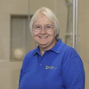 Freundliches Lächeln einer Frau in blauer Arbeitskleidung Eine lächelnde Frau mit Brille trägt ein blaues Polohemd und steht vor einer hellen, modernen Umgebung.