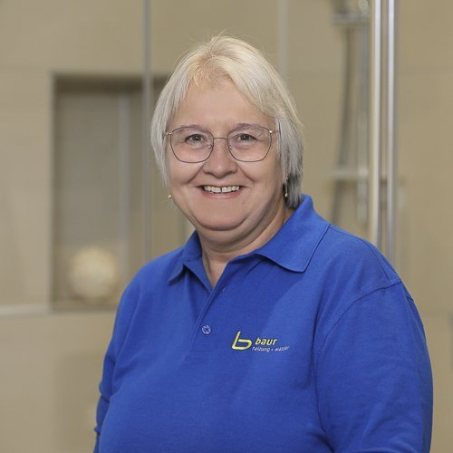 Eine lächelnde Frau mit Brille trägt ein blaues Polohemd und steht vor einer hellen, modernen Umgebung.