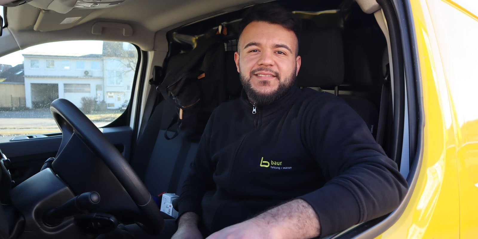 Der Fahrer in einem schwarzen Pullover zeigt ein freundliches Lächeln, während er in einem gelben Lieferwagen sitzt.