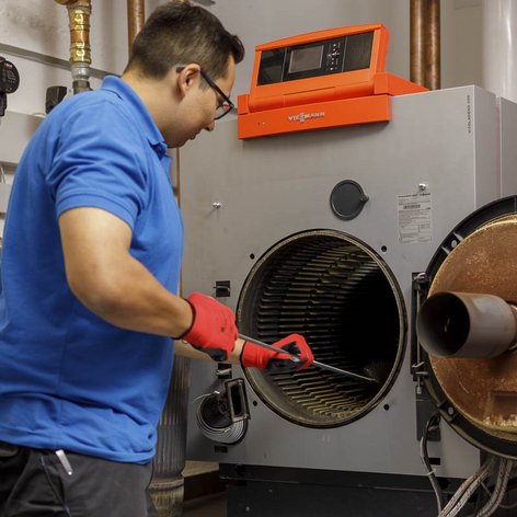 Ein Techniker in blauer Uniform reinigt eine Heizungsanlage mit einem Werkzeug in der Hand, während er konzentriert arbeitet.