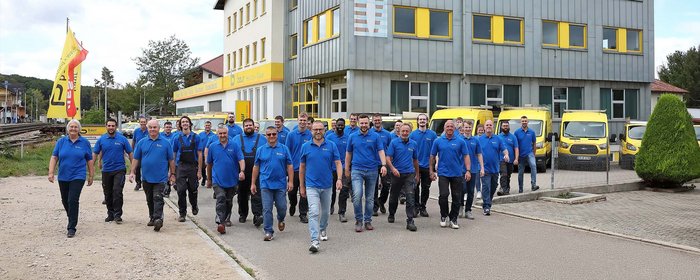 Eine Gruppe von Mitarbeitern in blauen Shirts geht selbstbewusst auf einem Weg vor einem Firmengebäude mit gelben Fahrzeugen.