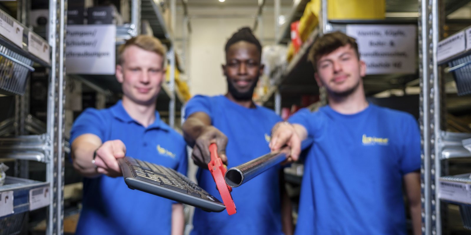 In einem Lager stehen drei Männer, die jeweils ein Werkzeug präsentieren: eine Tastatur, eine Zange und ein Metallrohr.