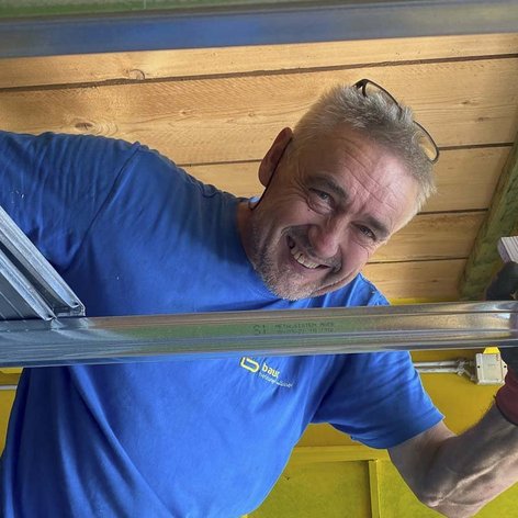 Ein Handwerker lächelt fröhlich, während er mit einem Metallregal in einem hellen Raum arbeitet, umgeben von Holz und gelben Wänden.