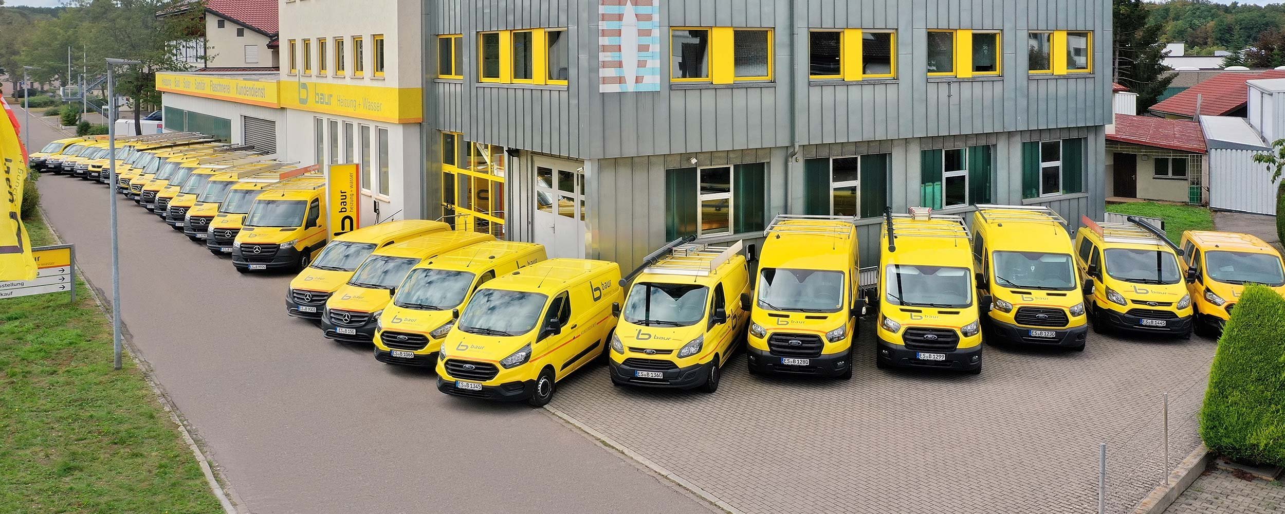 Eine Reihe von gelben Lieferwagen parkt ordentlich vor einem modernen Gebäude mit großen Fenstern und einem auffälligen Logo.