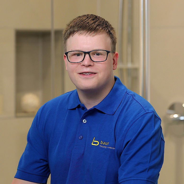 Der Junge trägt ein blaues Poloshirt mit dem Logo 'baur' und sitzt in einem hellen, modernen Raum mit sanften Farben.
