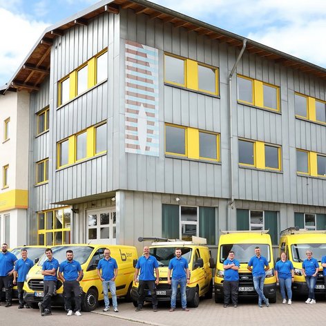 Eine Gruppe von Mitarbeitern in blauen T-Shirts steht vor einem modernen Gebäude mit gelben Fenstern und Lieferwagen.