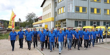 Eine Gruppe von Mitarbeitern in blauen Hemden geht selbstbewusst auf einem Gehweg vor einem modernen Gebäude mit gelben Fenstern.
