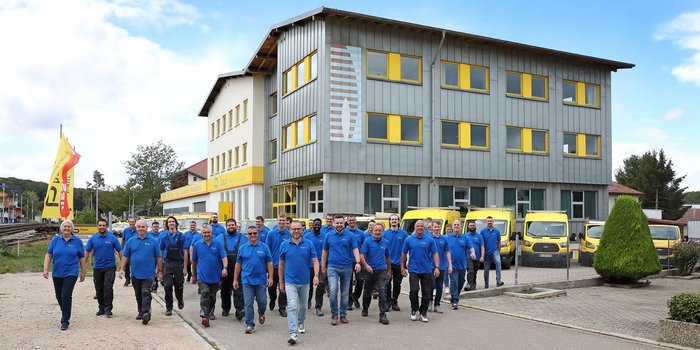Eine Gruppe von Mitarbeitern in blauen Hemden geht selbstbewusst auf einem Gehweg vor einem modernen Gebäude mit gelben Fenstern.