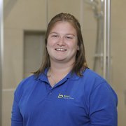 Freundliches Porträt einer Frau in professioneller Umgebung Eine lächelnde Frau mit langen, braunen Haaren trägt ein blaues Polohemd und steht in einem modernen, hellen Raum.