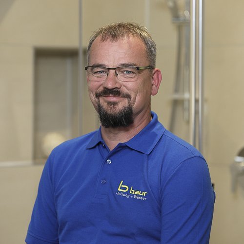 Ein lächelnder Mann mit Brille trägt ein blaues Poloshirt und steht vor einer modernen Badezimmerumgebung.