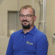 Professioneller Handwerker in blauer Arbeitskleidung lächelt freundlich Ein lächelnder Mann mit Brille trägt ein blaues Poloshirt und steht vor einer modernen Badezimmerumgebung.