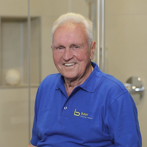 Ein älterer Mann mit einem freundlichen Lächeln trägt ein blaues Polohemd mit dem Logo 'baur' und sitzt in einem modernen Raum.