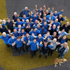 Eine große Gruppe von Menschen in blauen T-Shirts steht auf einer Straße und winkt fröhlich in die Kamera.