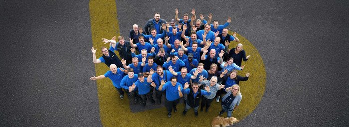 Eine große Gruppe von Menschen in blauen T-Shirts steht auf einer Straße und winkt fröhlich in die Kamera.