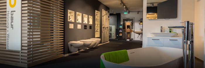 Der Ausstellungsraum zeigt eine freistehende Badewanne mit einem grünen Handtuch, umgeben von verschiedenen Toiletten und Waschbecken.