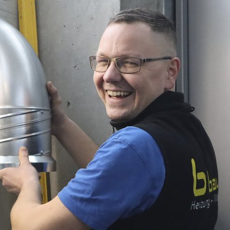 Ein lächelnder Mann hält ein silbernes Rohr in einer industriellen Umgebung, während er mit Freude arbeitet.