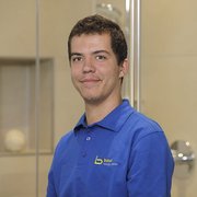 Junger Mann in blauer Arbeitskleidung lächelt freundlich Ein junger Mann mit kurzem, braunem Haar trägt ein blaues Polohemd mit dem Logo von Baur und lächelt freundlich.