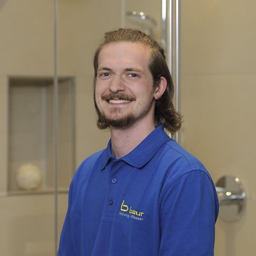 Ein junger Mann mit langen Haaren lächelt freundlich in einem modernen Badezimmer mit Glasduschkabine im Hintergrund.
