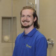 Professioneller Handwerker in blauer Arbeitskleidung lächelt freundlich Ein junger Mann mit langen Haaren lächelt freundlich in einem modernen Badezimmer mit Glasduschkabine im Hintergrund.