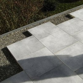 Eine moderne Terrasse mit grauen Platten, die auf Kies verlegt sind, umgeben von üppigem Grün und Sträuchern.